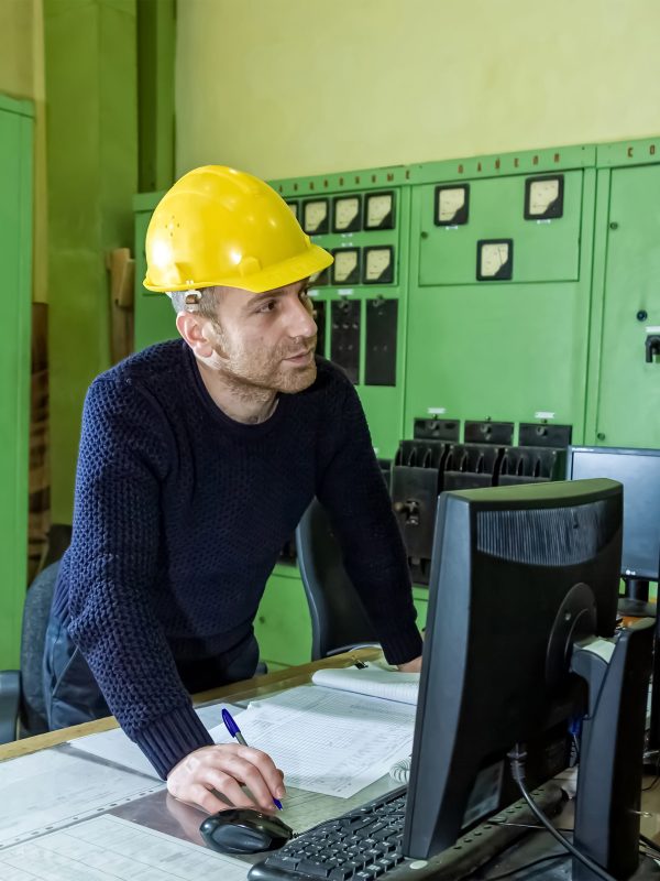 A vertical shot of a factory employee wearing a yellow helmet working on his computer